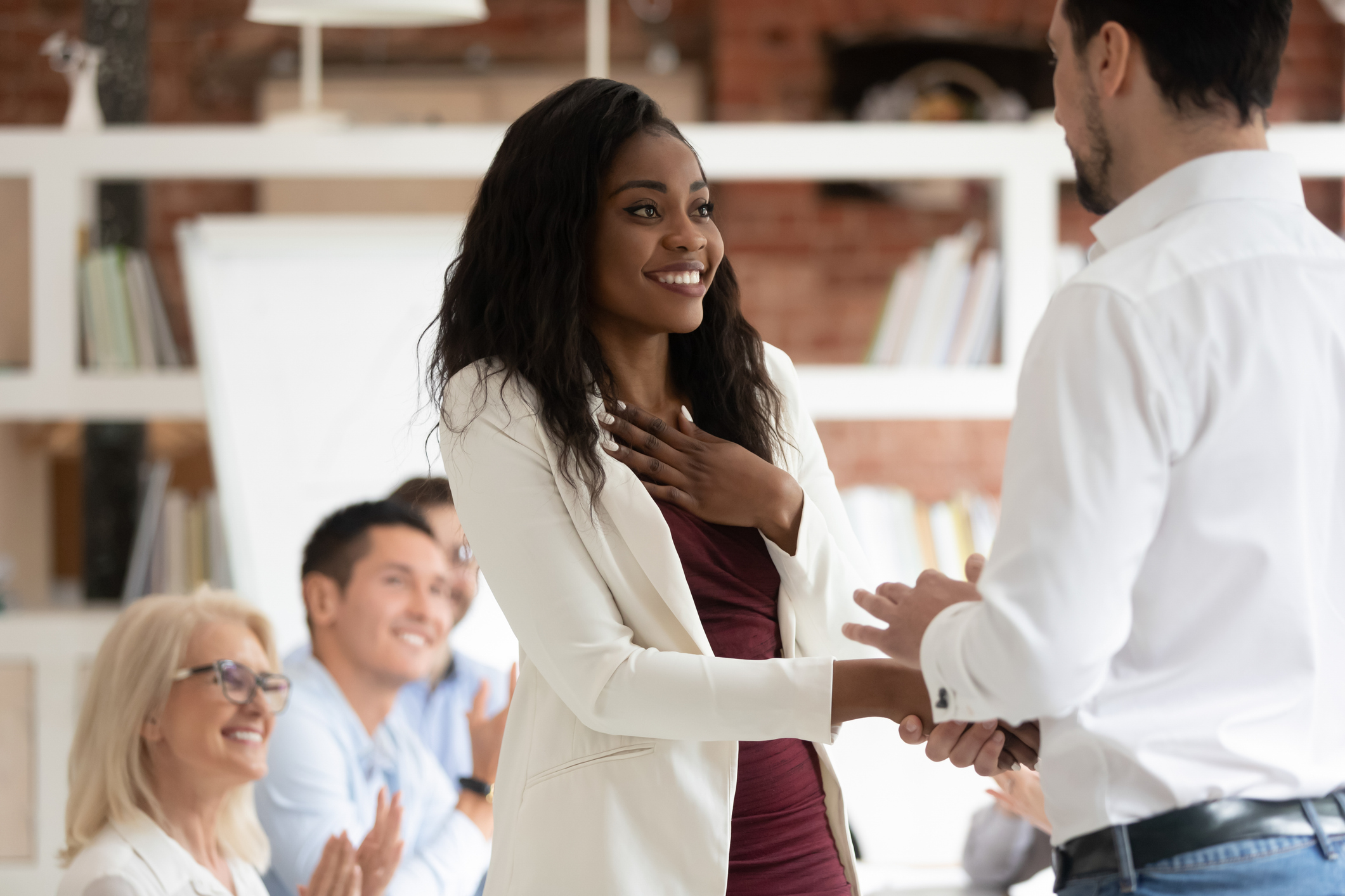 Photo of happy black female employee getting rewarded for professional achievement and having a handshake with boss. Happy employees in background.