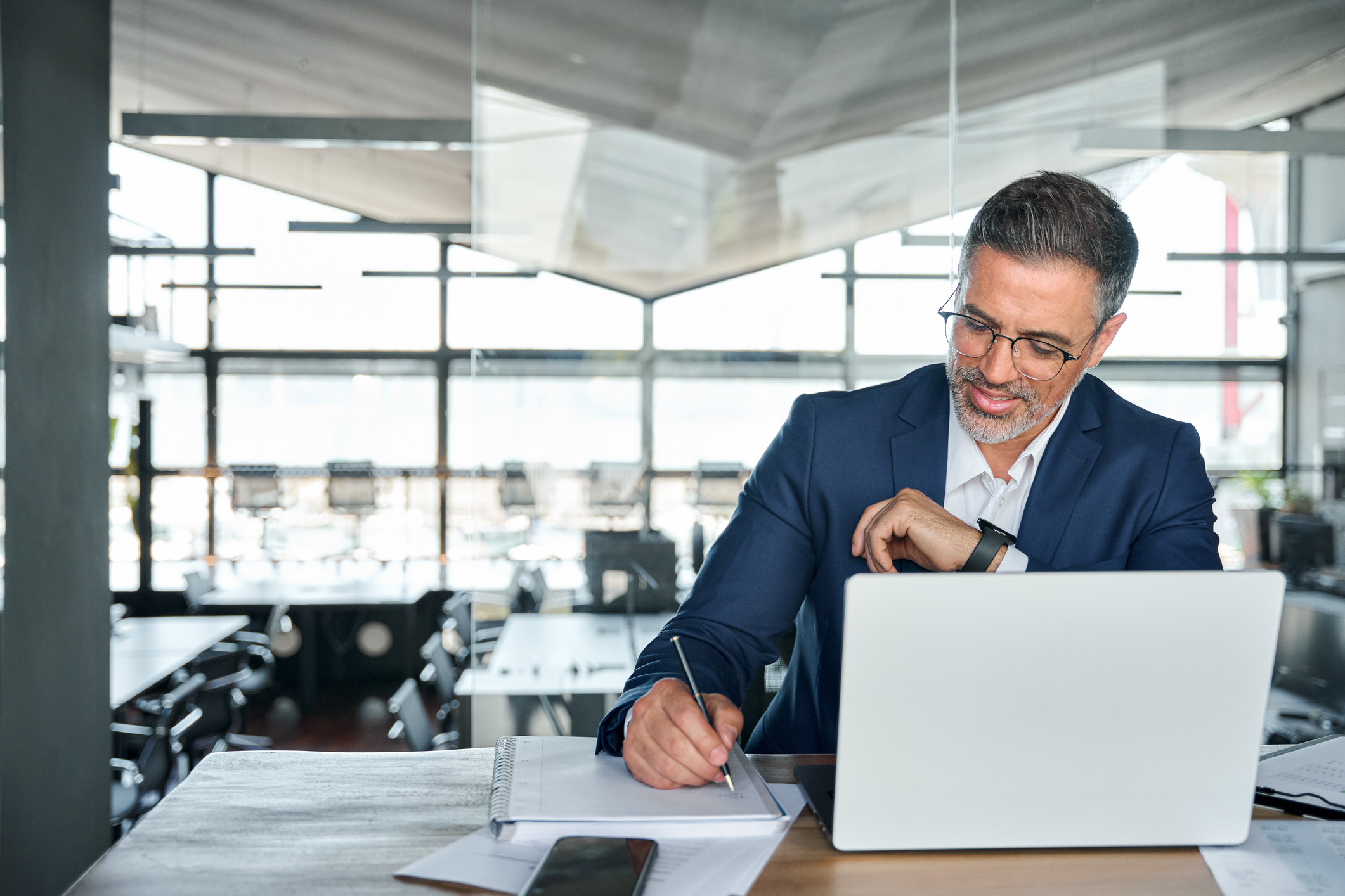 Photo of mid aged business man working on laptop computer in office writing notes.